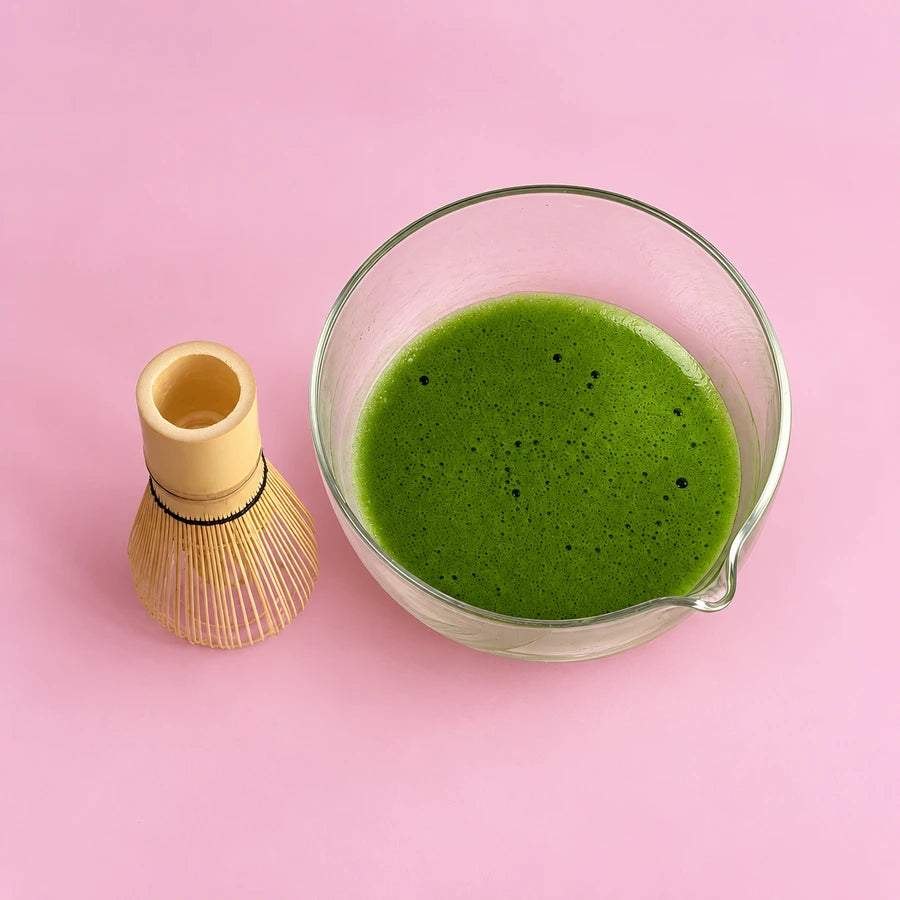 A bamboo whisk beside freshly whisked matcha in a glass pouring bowl on pink