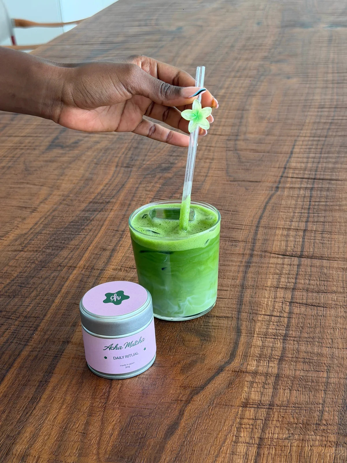 A hand stirring an iced matcha beside an Acha Matcha tin