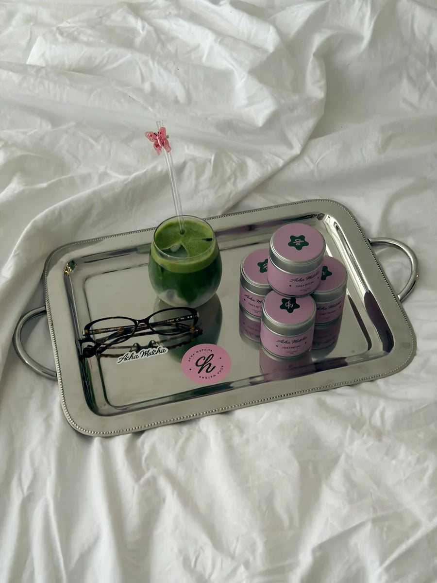 A silver tray of Acha Matcha tins and an iced matcha on white bedding